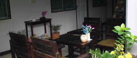 A wooden table and chairs set up in a room with a white wall and green window bars. There are potted plants and a small vase with flowers on the table.