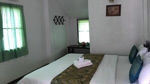 A hotel room with a bed, a window with green curtains, an air conditioning unit, a small table with a vase of flowers, and a wall-mounted light fixture. - Ampawan Guesthouse (Chiang Mai)