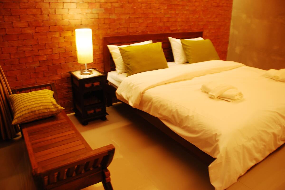 A hotel room with a brick wall, a wooden bench, a nightstand with a lamp, a bed with white bedding, and a green pillow.