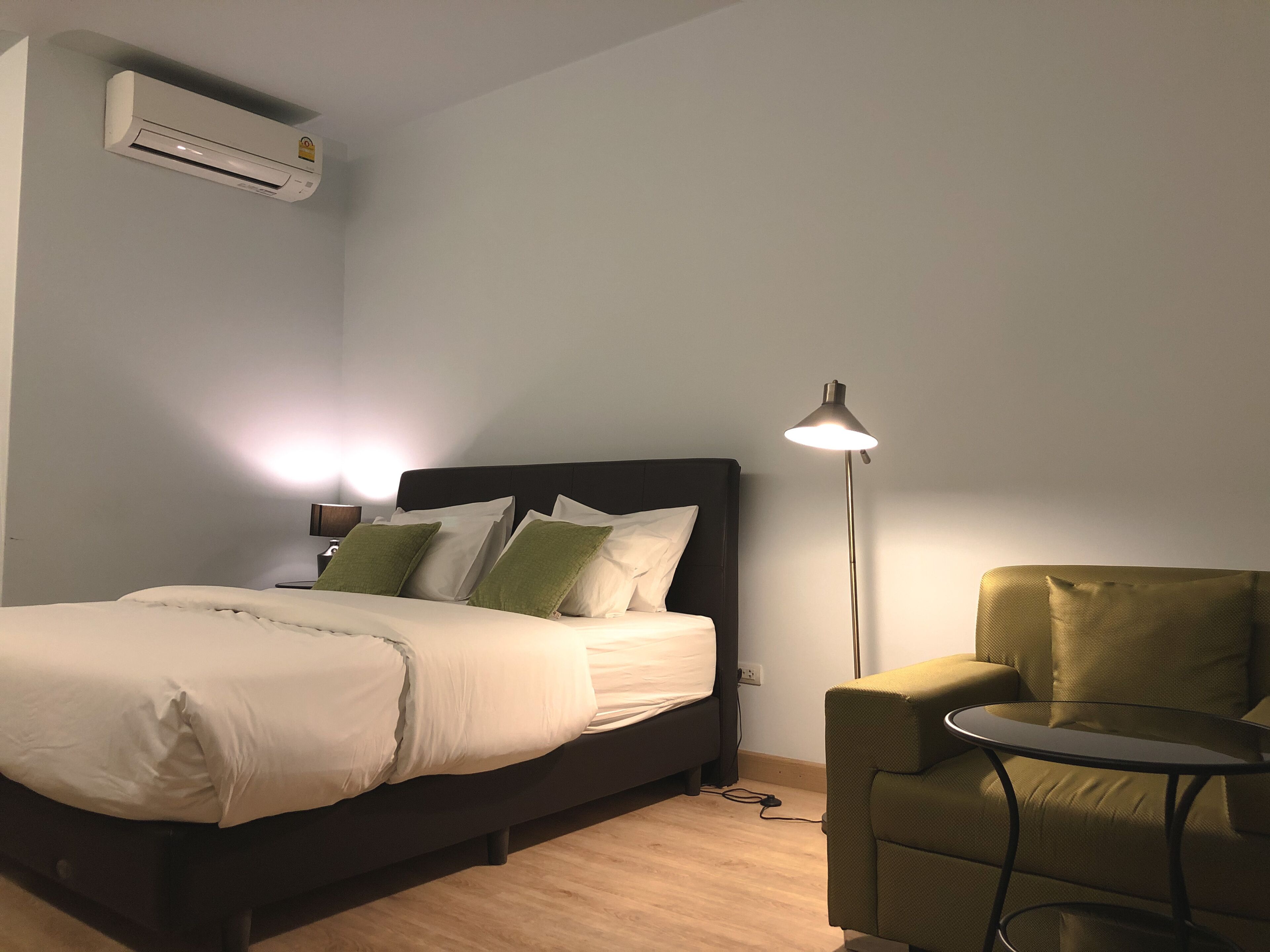 A neatly made bed with white bedding and green pillows, a bedside lamp, a small round table, and a green armchair.