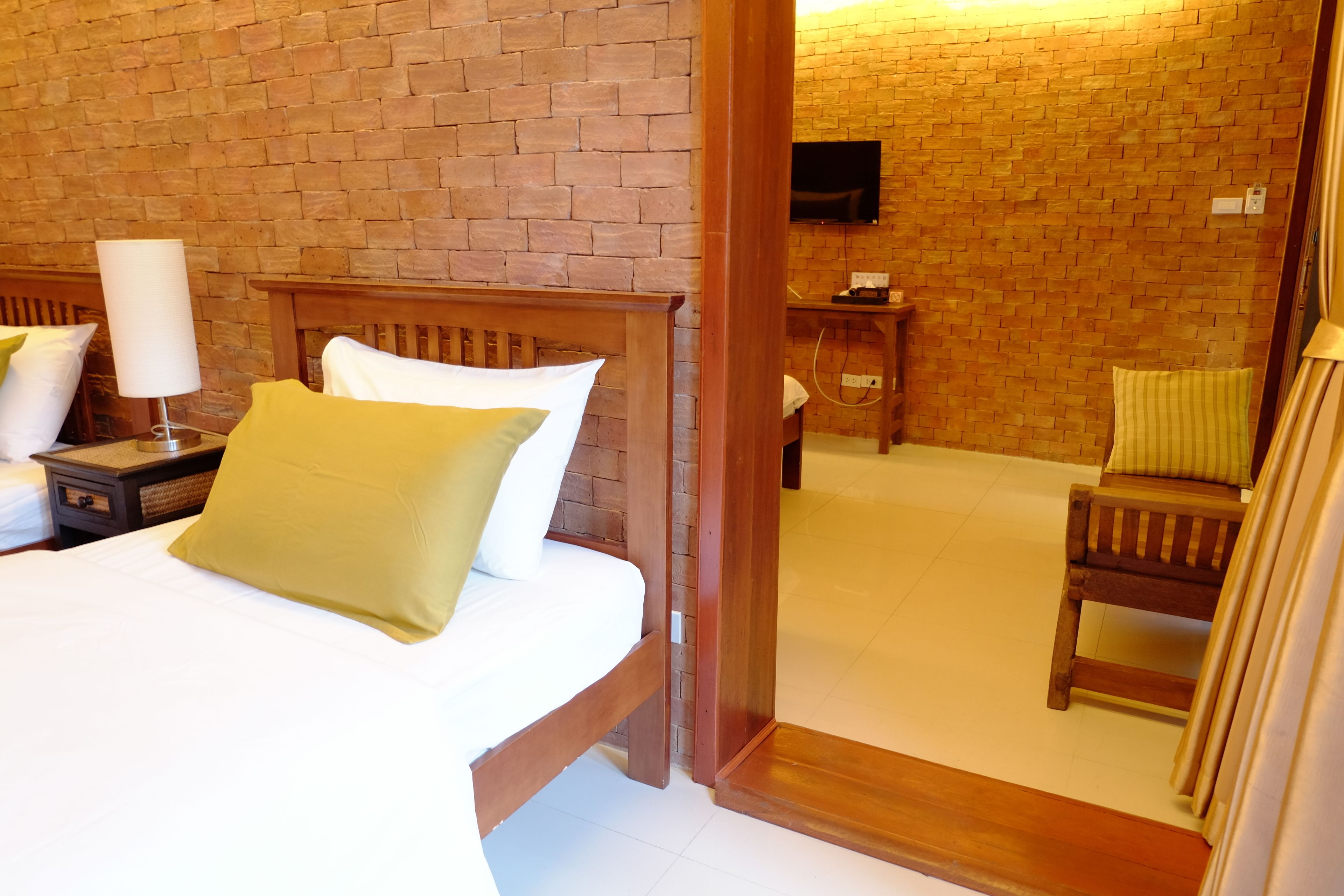 A hotel room with a brick wall, a wooden bed, a bedside table with a lamp, a chair, and a television mounted on the wall.
