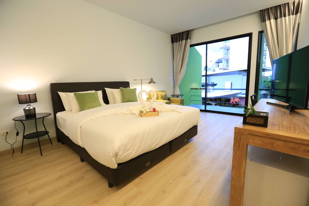A hotel room with a large bed, a bedside table with a lamp, a small table with a plant, and a view of the outside through a large window.