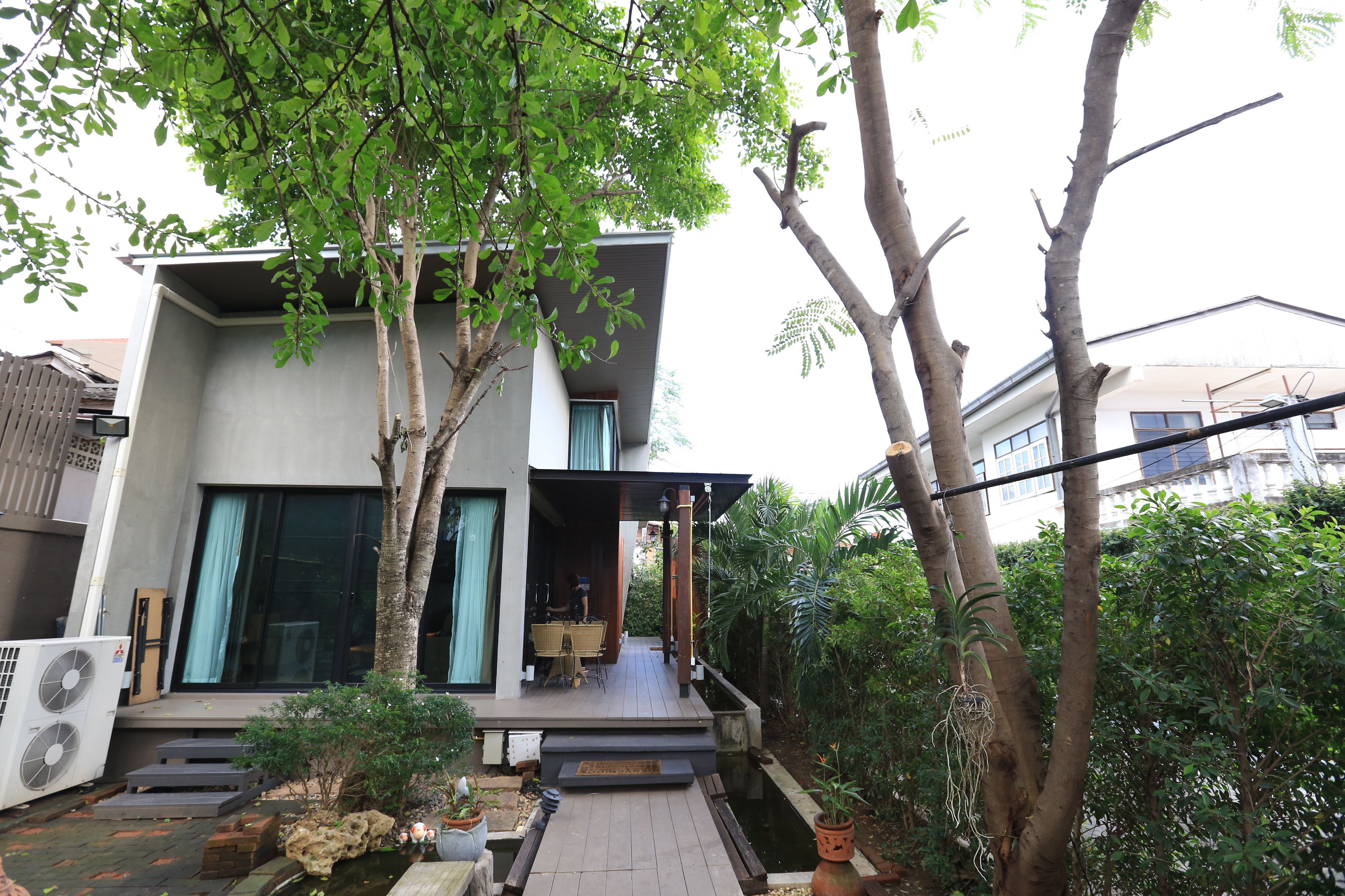 A modern two-story house with a covered porch, a small garden area with plants and a water feature, and a large air conditioning unit on the ground floor.