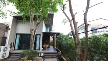 A modern two-story house with a covered porch, a small garden area with plants and a water feature, and a large air conditioning unit on the ground floor.