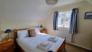 A neatly made bed with white linens and folded towels, a wooden headboard, a colorful decorative pillow, a bedside lamp, and a window with blue curtains. - Walma has become a firm favourite with holidaymakers , a perfect retreat for family and friends. (Cromer)