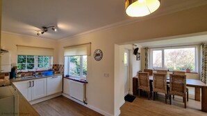 A kitchen with white cabinets, a sink, and a window with a view of greenery. There is a dining area with wicker chairs and a table. - Walma has become a firm favourite with holidaymakers , a perfect retreat for family and friends. (Cromer)