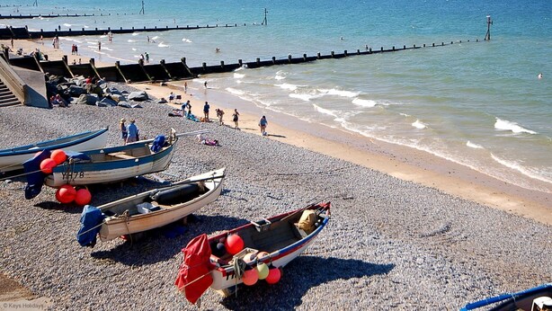 Una playa pedregosa con varias embarcaciones pequeñas, un muelle y gente disfrutando del paseo marítimo.