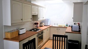 Cocina compacta con armarios blancos, encimera de madera, estufa, lavabo y un pequeño comedor con una silla.