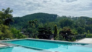 A swimming pool with a view of a forested hill and a building in the background. - Hotel Alta Vista (Ella)