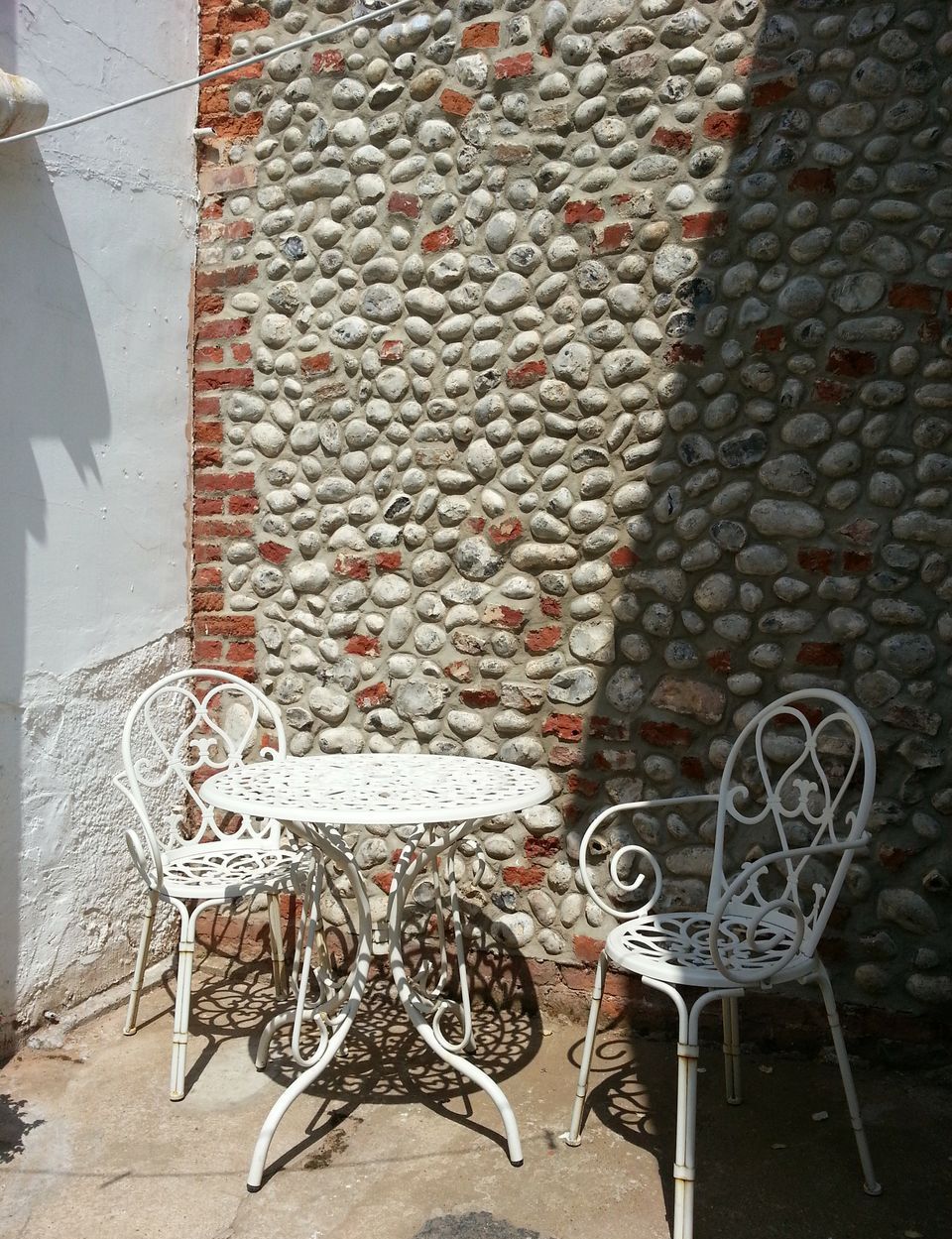 Un pequeño espacio al aire libre con una mesa y sillas de metal blanco, frente a una pared texturizada de piedra y ladrillo.