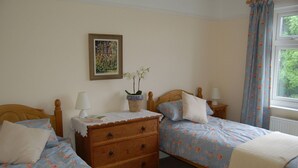 A bedroom with two single beds, a wooden dresser, a bedside table with a lamp, a framed picture on the wall, and a window with curtains. - This delightful semi-detached cottage is close to walks,  bird watching & minutes from the beach. (Sheringham)