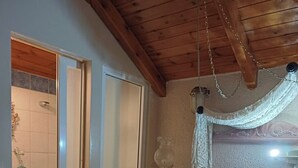 A bedroom with a wooden ceiling, a bed with white linens and a floral duvet, a nightstand with a lamp, and a bathroom with a shower and toilet. - Family Abode for Vacation in Arachova (Distomo-Arachova-Antikyra)