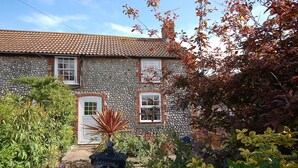 A stone building with a tiled roof, a white door, and a small garden with various plants and flowers. - Romany Rye is one of the oldest and prettiest Fisherman's cottage in Sheringham in a great location (Sheringham)