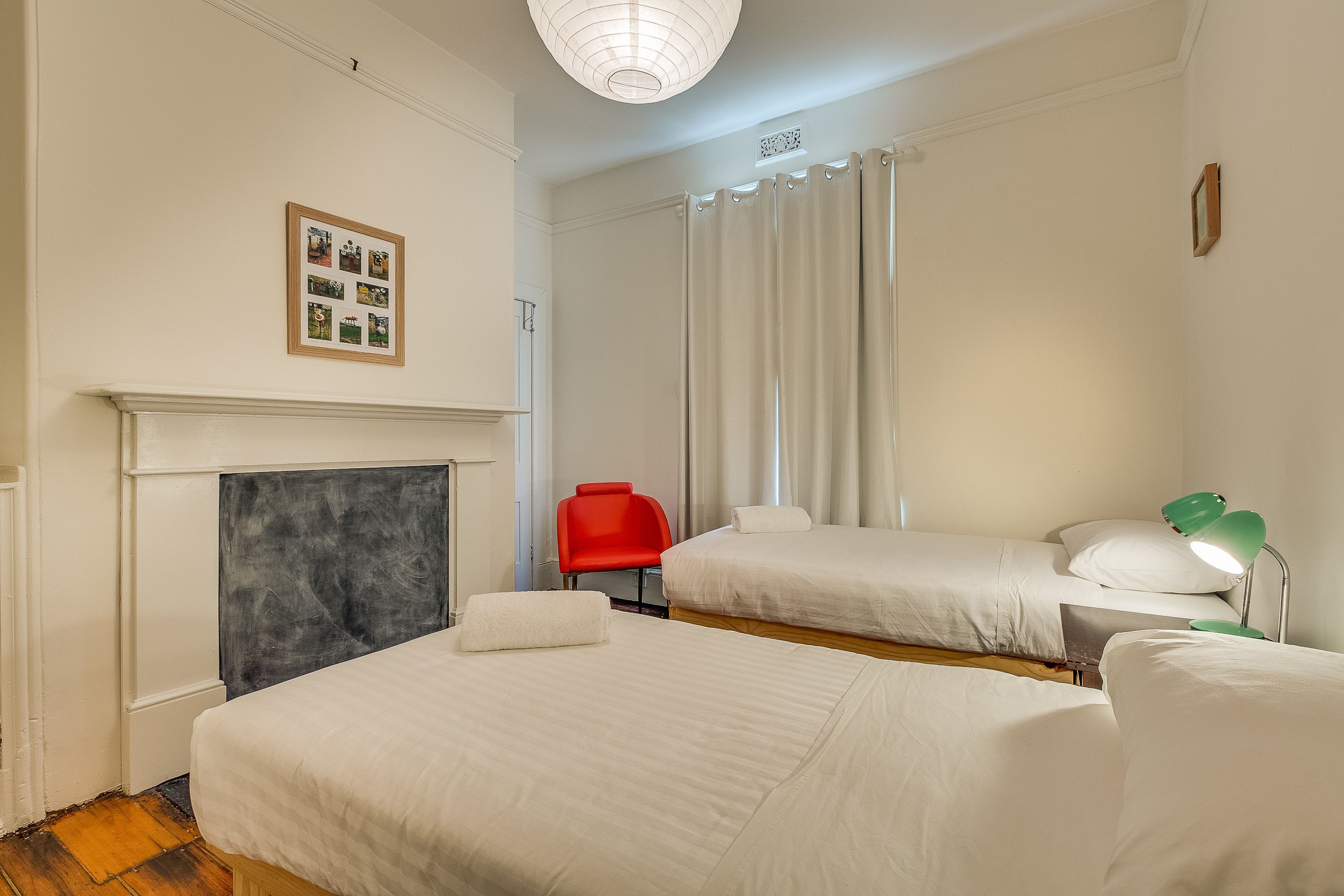 A hotel room with two beds, a red chair, a green desk lamp, and a framed photo on the wall.