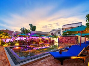 A pool area with lounge chairs and umbrellas, surrounded by buildings and palm trees. - Lilly Holiday Hotel (Siem Reap)