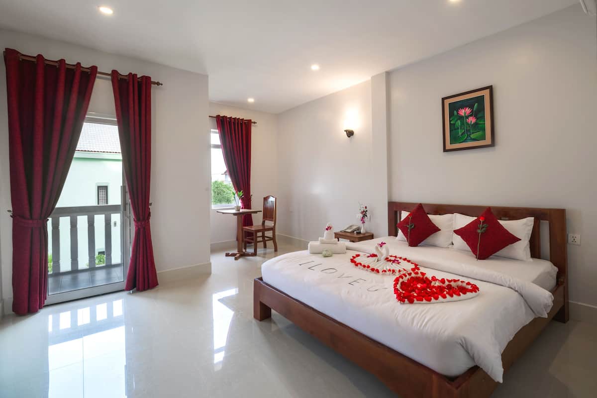 Habitación de hotel con una cama grande, una mesita pequeña con una silla y un balcón con cortinas rojas.