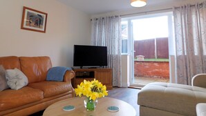 Una sala de estar con un sofá de cuero marrón, un sillón beige, una mesa de centro con un jarrón de flores amarillas, un televisor de pantalla plana sobre un mueble de madera y un ventanal corredero que da a un jardín.