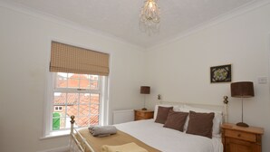 Ein Schlafzimmer mit einem großen Bett, einem Fenster mit Vorhängen in Karomuster, einem Heizkörper, einem Nachttisch mit Lampe und einem Bild an der Wand.