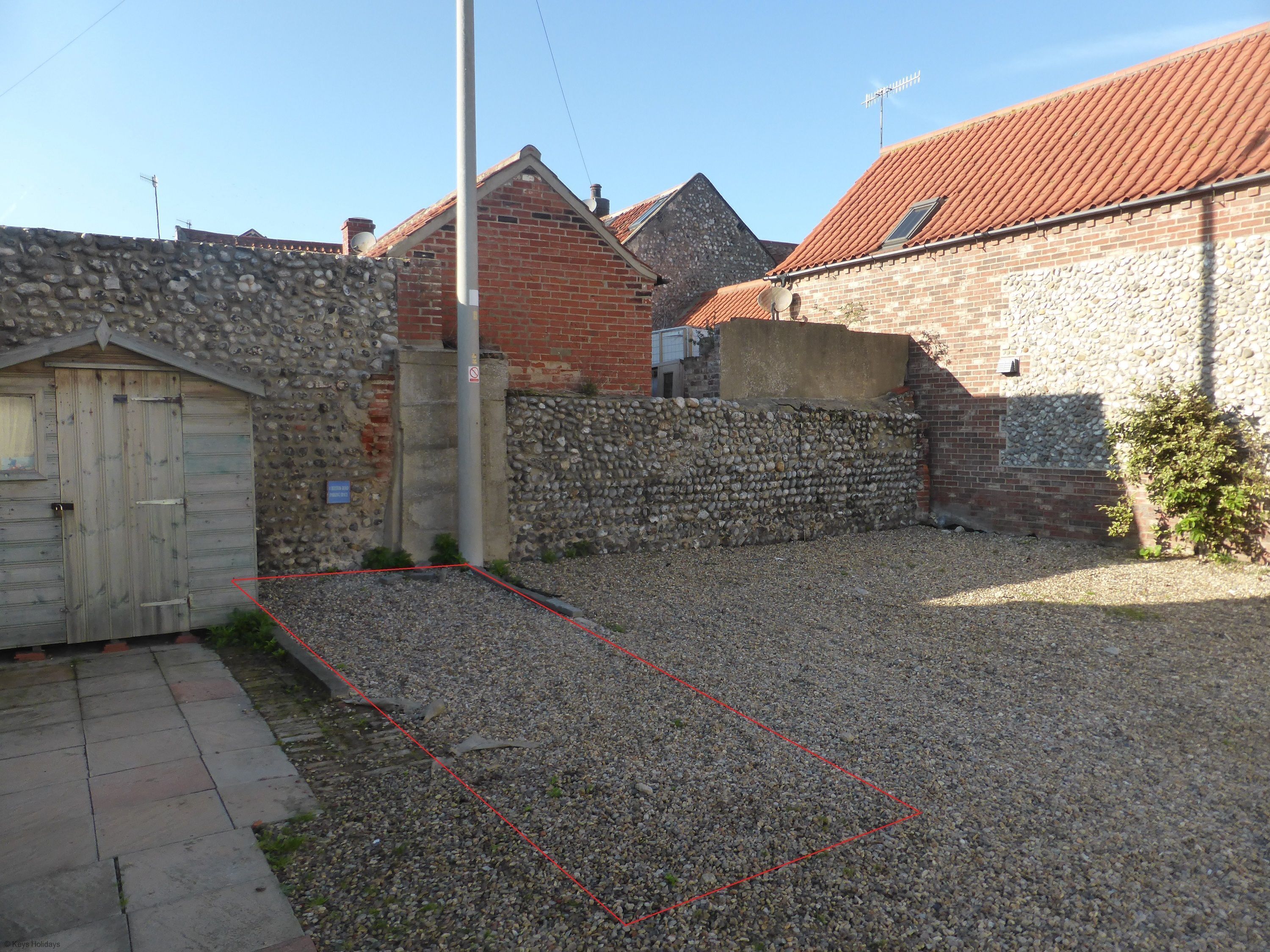 Une allée de gravier mène à un mur de pierres surmonté d’un portail en bois, puis à un bâtiment en briques avec un toit recouvert de tuiles rouges, et enfin à un petit jardin.