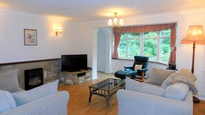 A living room with a stone fireplace, a flat-screen TV, a comfortable sofa, a glass coffee table, and a view of greenery through a window. - Set within an area of Outstanding Natural Beauty this bungalow is just the place in which to relax (Cromer)