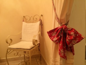A metal-framed chair with a white cushion, positioned next to a wall with a white curtain and a red floral cloth tied to it.