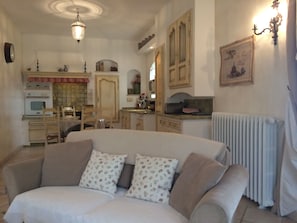 A cozy living room with a sofa, a dining table, and chairs. There is a kitchen area with cabinets and a radiator.