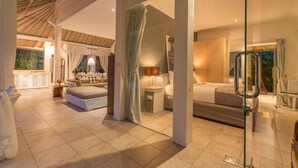 A spacious bedroom with a large bed, a sitting area with a sofa and armchairs, a glass partition, and a view of the outdoors. - Villa Esha Drupadi I (Seminyak)