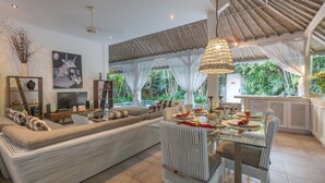 A modern living room with a sofa, dining table, and chairs. There is a TV, a bookshelf, and a large window with curtains. - Villa Esha Drupadi I (Seminyak)