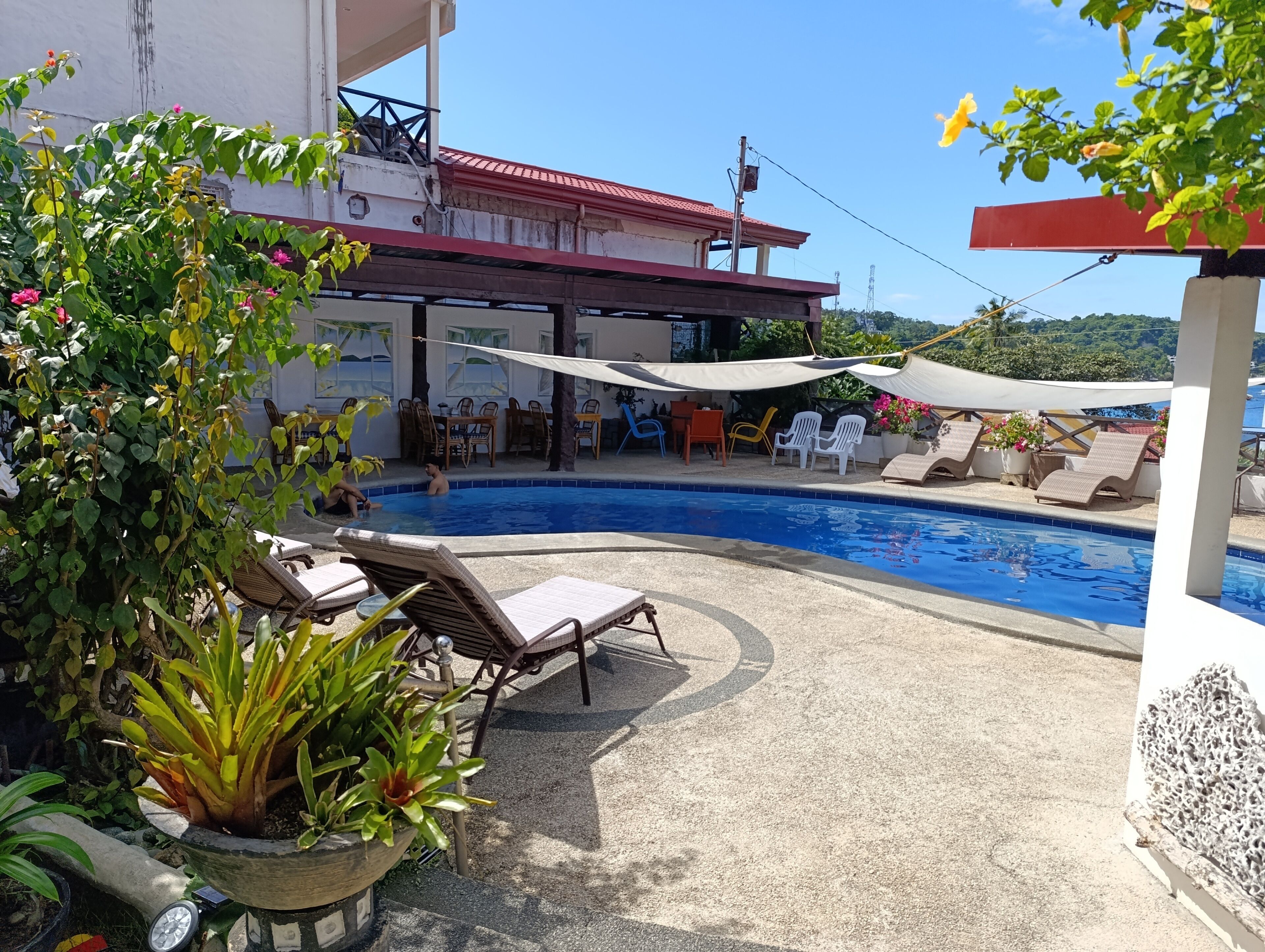 A swimming pool with lounge chairs and umbrellas, surrounded by potted plants and a building with a red roof.
