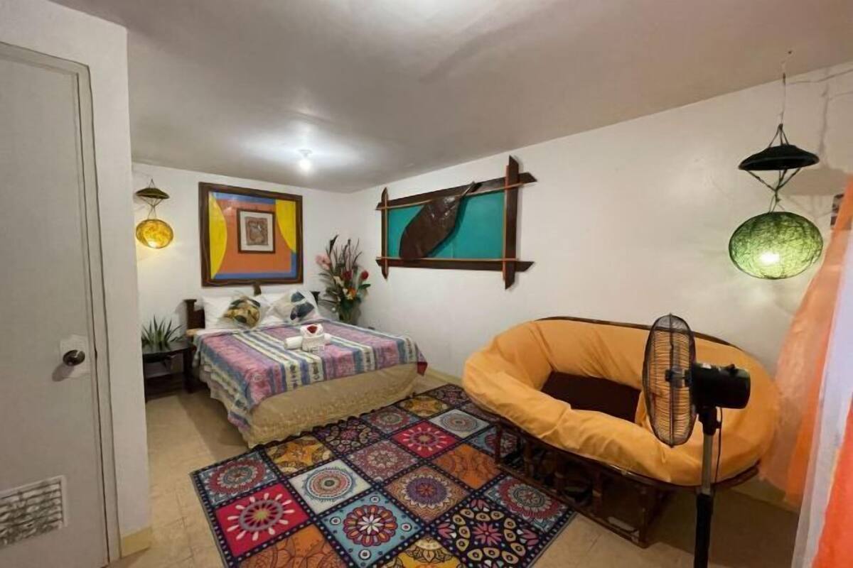A bedroom with a bed, a chair, a fan, and artwork on the walls.