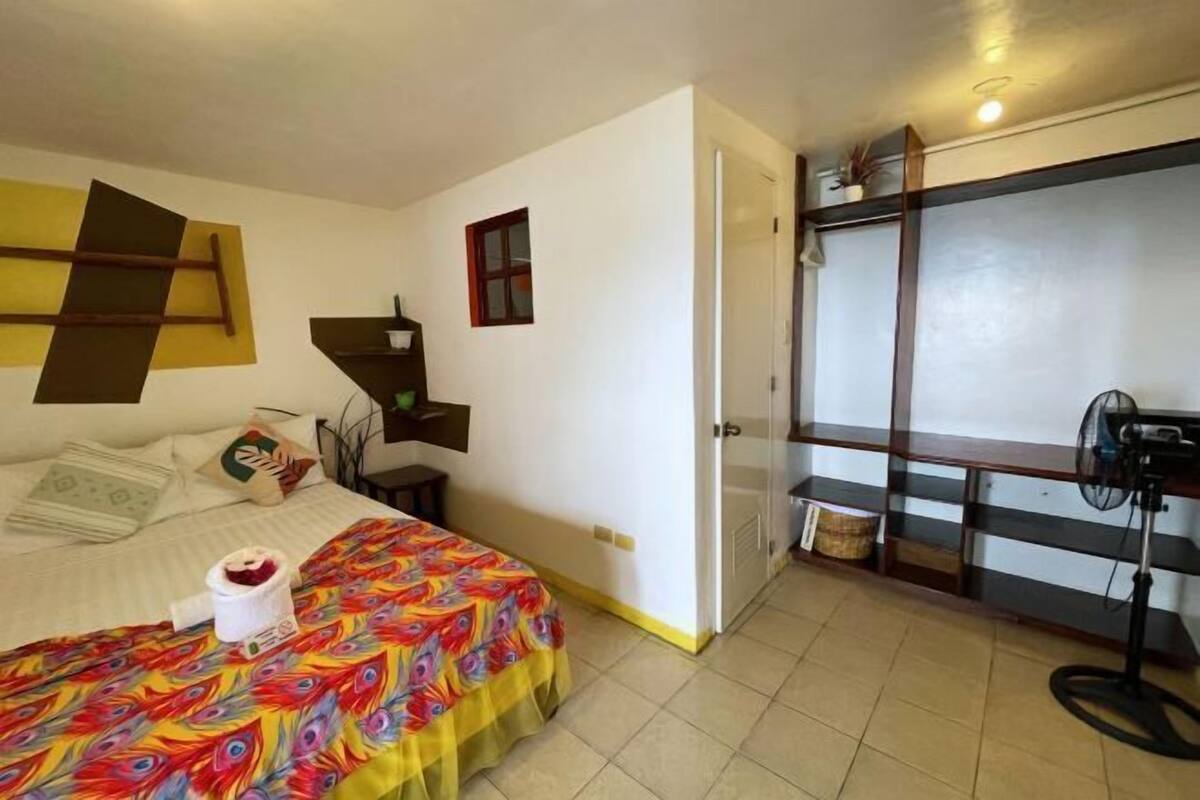 A bedroom with a bed, a desk, a chair, a fan, and a wardrobe.