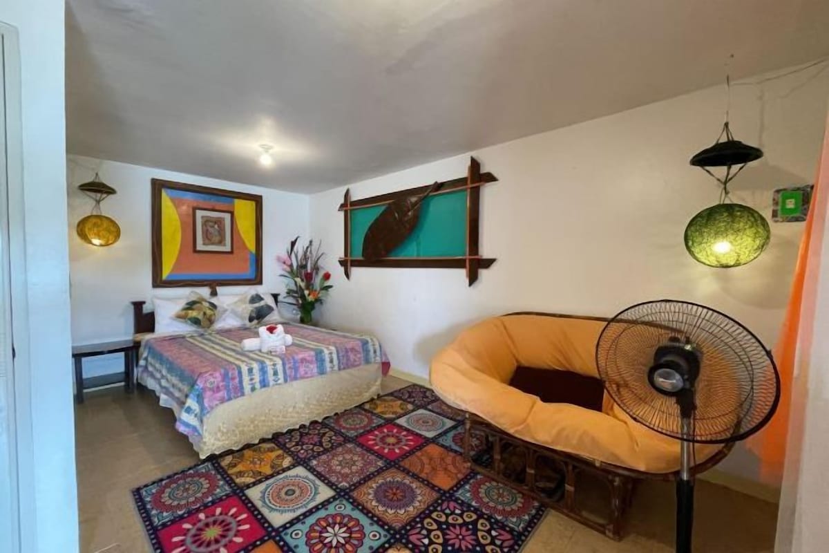 Une chambre à coucher avec un lit, une chaise, un ventilateur sur pied et un tapis à motifs.