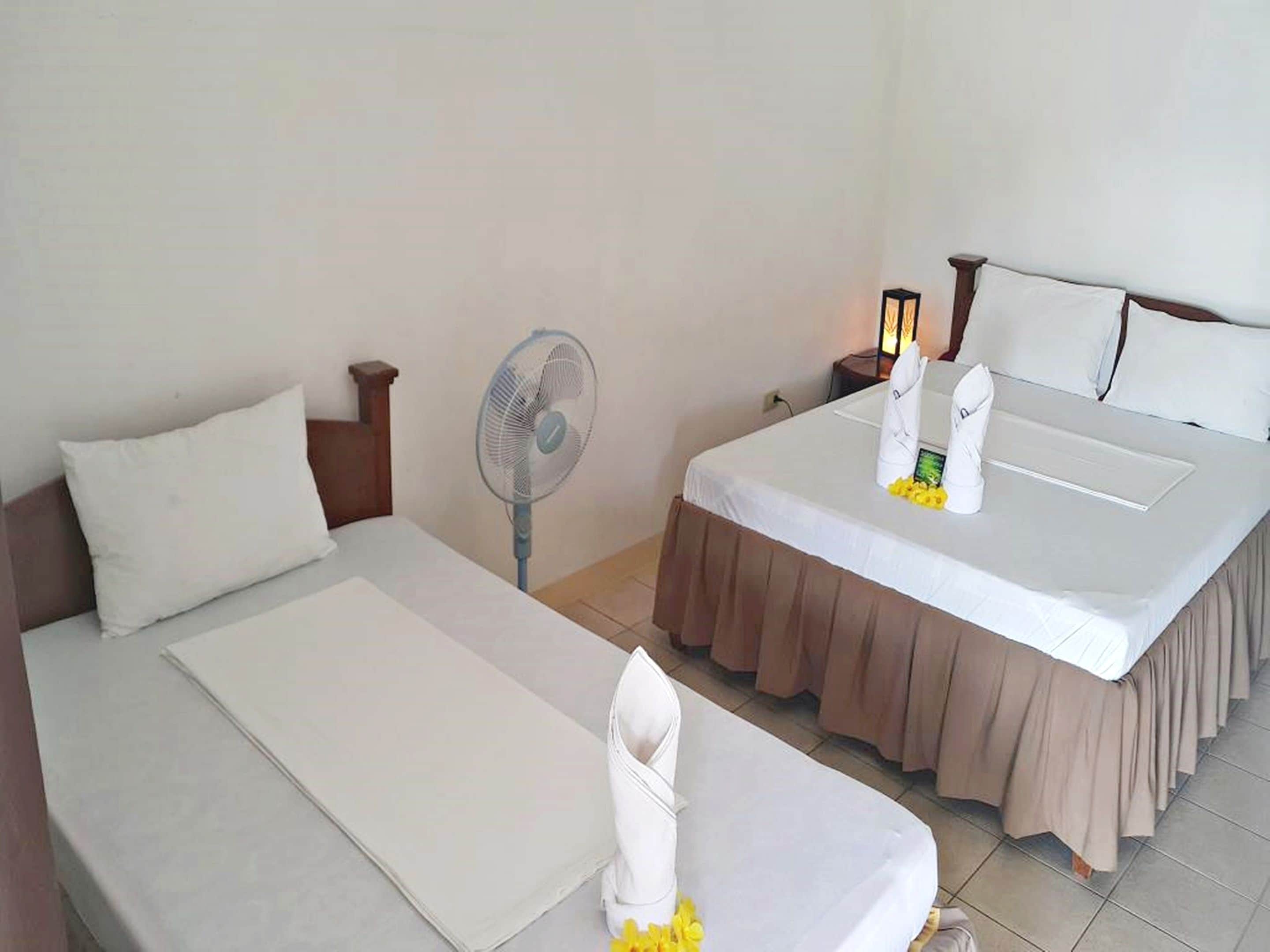 A hotel room with two beds, a standing fan, a nightstand with a lamp, and a towel on the bed.