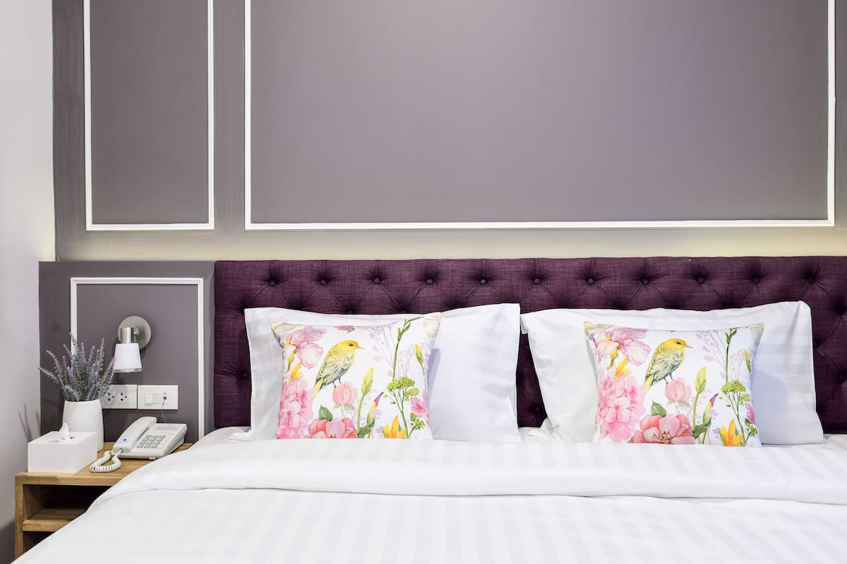 Un lit bien fait, avec du linge de lit blanc, une tête de lit violette à motifs capitonnés et deux coussins décoratifs ornés de motifs floraux.