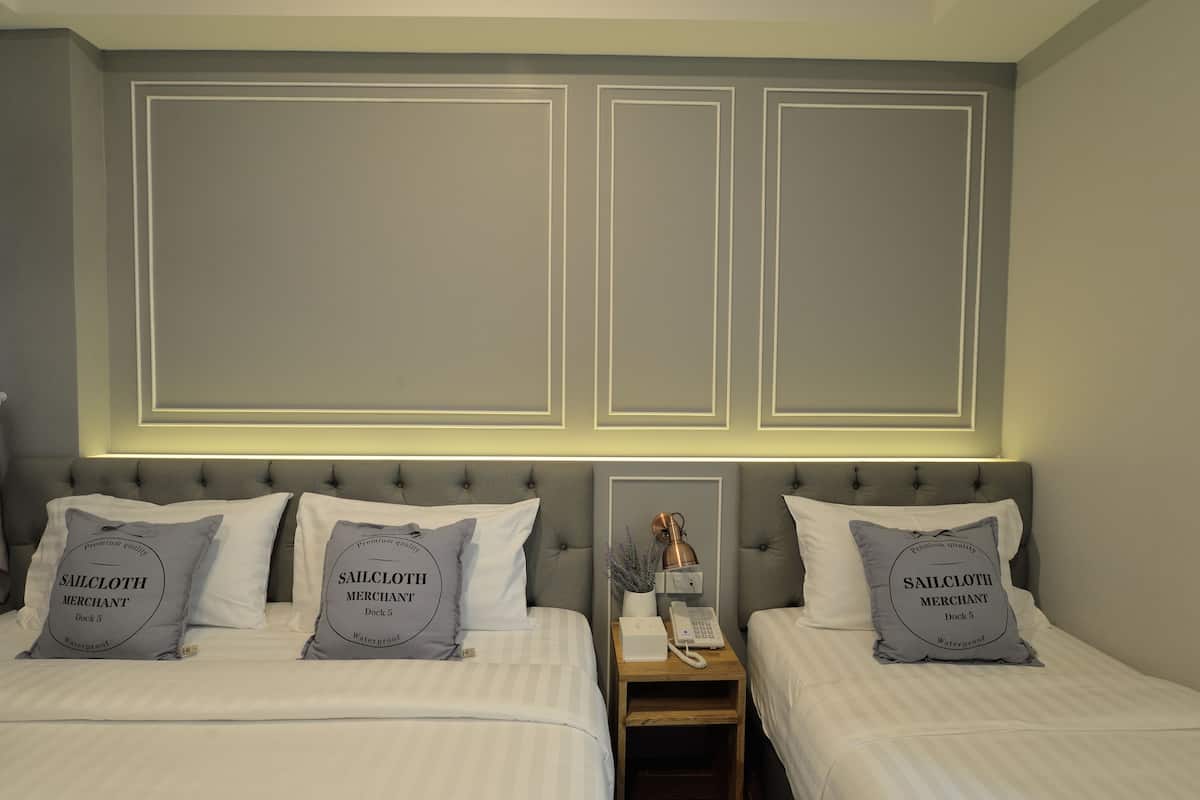 Une chambre d’hôtel avec deux lits, chacun doté d’une tête de lit grise et d’oreillers portant l’inscription « Sailcloth Merchant Dock 5 Waterproof ». Il y a une table de chevet avec une lampe, un téléphone et un vase de fleurs placé entre les deux lits.