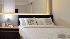 A neatly made bed with white linens and floral pillows, a bedside table with a lamp, and a large mirror above the bed.