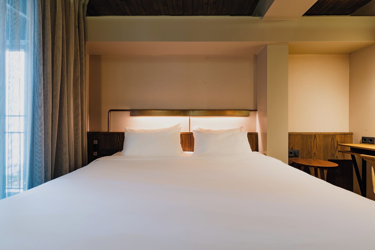 Une chambre d’hôtel avec un grand lit, une table de chevet, une petite table et une fenêtre avec des rideaux.