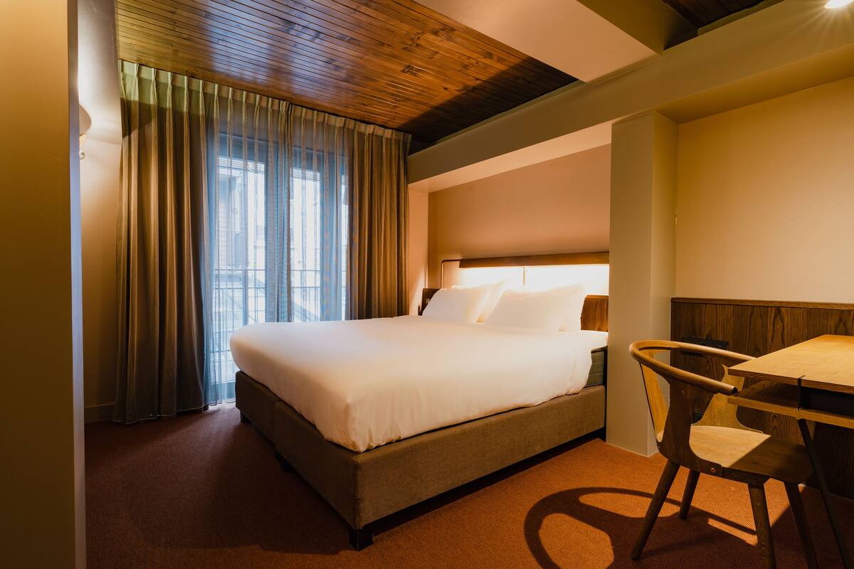Una habitación de hotel con una cama grande, techo de madera, ventana con cortinas y una mesita con una silla.