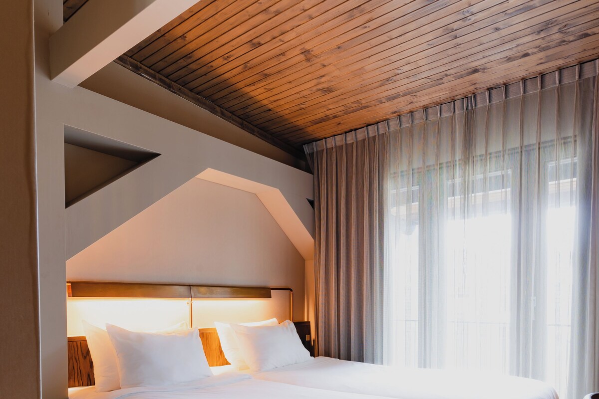 A modern hotel room with a large bed, wooden ceiling, and a view of the outdoors through sheer curtains.