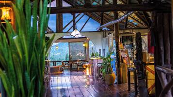 Un interior rústico con vigas de madera, un ventanal con vistas a un paisaje montañoso y una zona de estar con mesas y sillas.