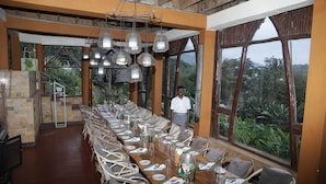 A long dining table set for a meal with wicker chairs, large windows offering a view of greenery, and hanging light fixtures.