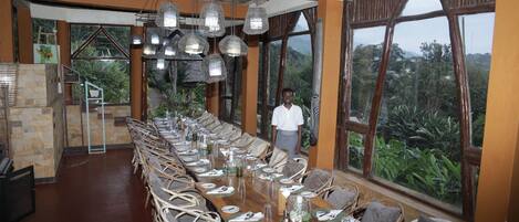 Una larga mesa de comedor lista para una comida, con sillas de mimbre, ventanas amplias que ofrecen vistas a la vegetación y lámparas colgantes.