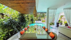 Una mesa de comedor con cubierta de vidrio, sillas de mimbre y cojines anaranjados, rodeada de abundante follaje y vistas a una piscina.