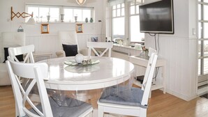 Un comedor con una mesa redonda, sillas blancas, un televisor montado en la pared y ventanas amplias.