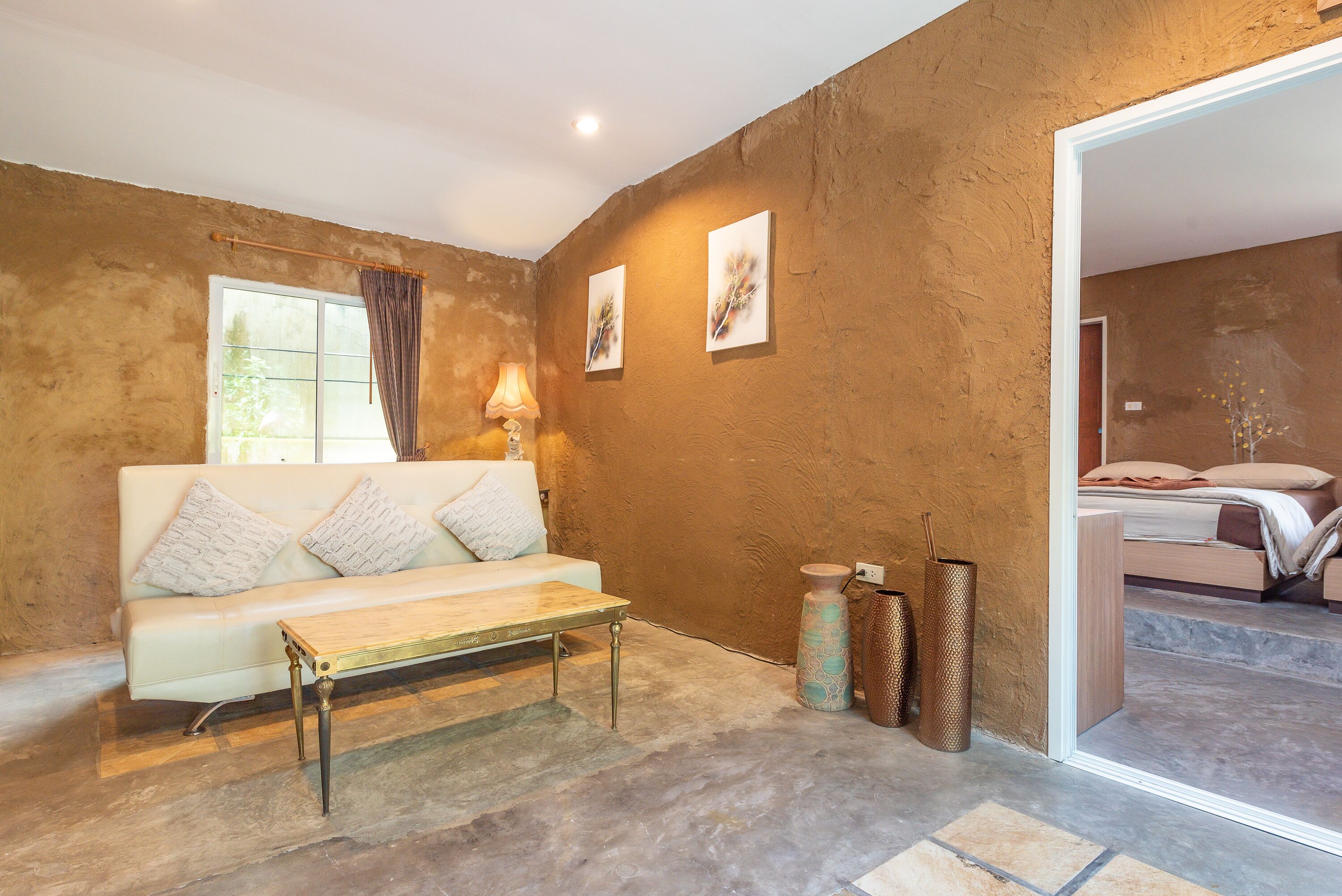 A room with a white sofa, a wooden coffee table, and a large window with curtains. There are two wall-mounted lamps and two framed artworks.