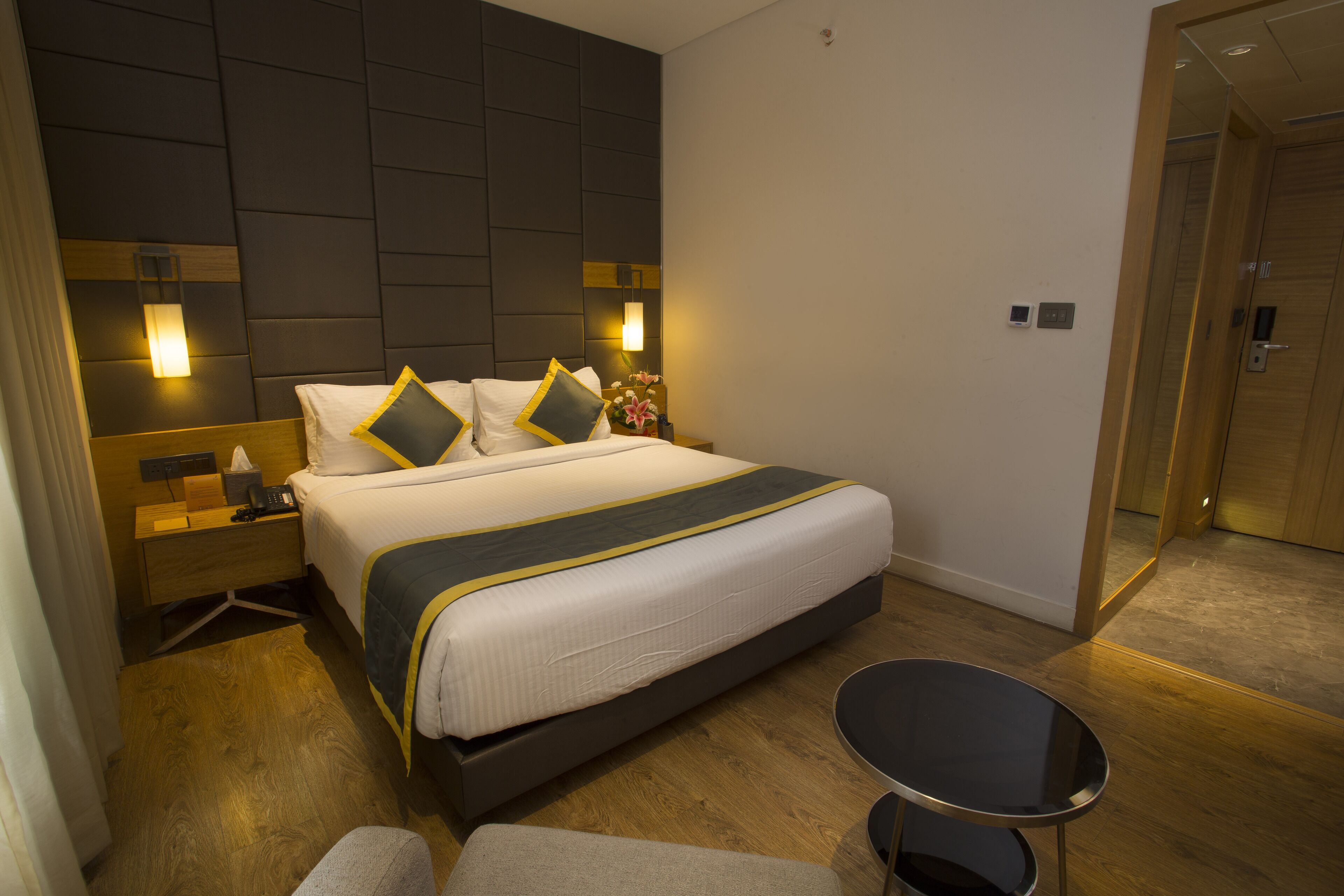 A modern hotel room with a large bed, bedside tables, a small round table, and a mirror.