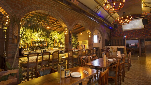 A restaurant with brick arches, wooden tables, and chairs. There is a bar area with shelves of bottles and a screen displaying a presentation. - Fountain Tree by TGI (Bengaluru)