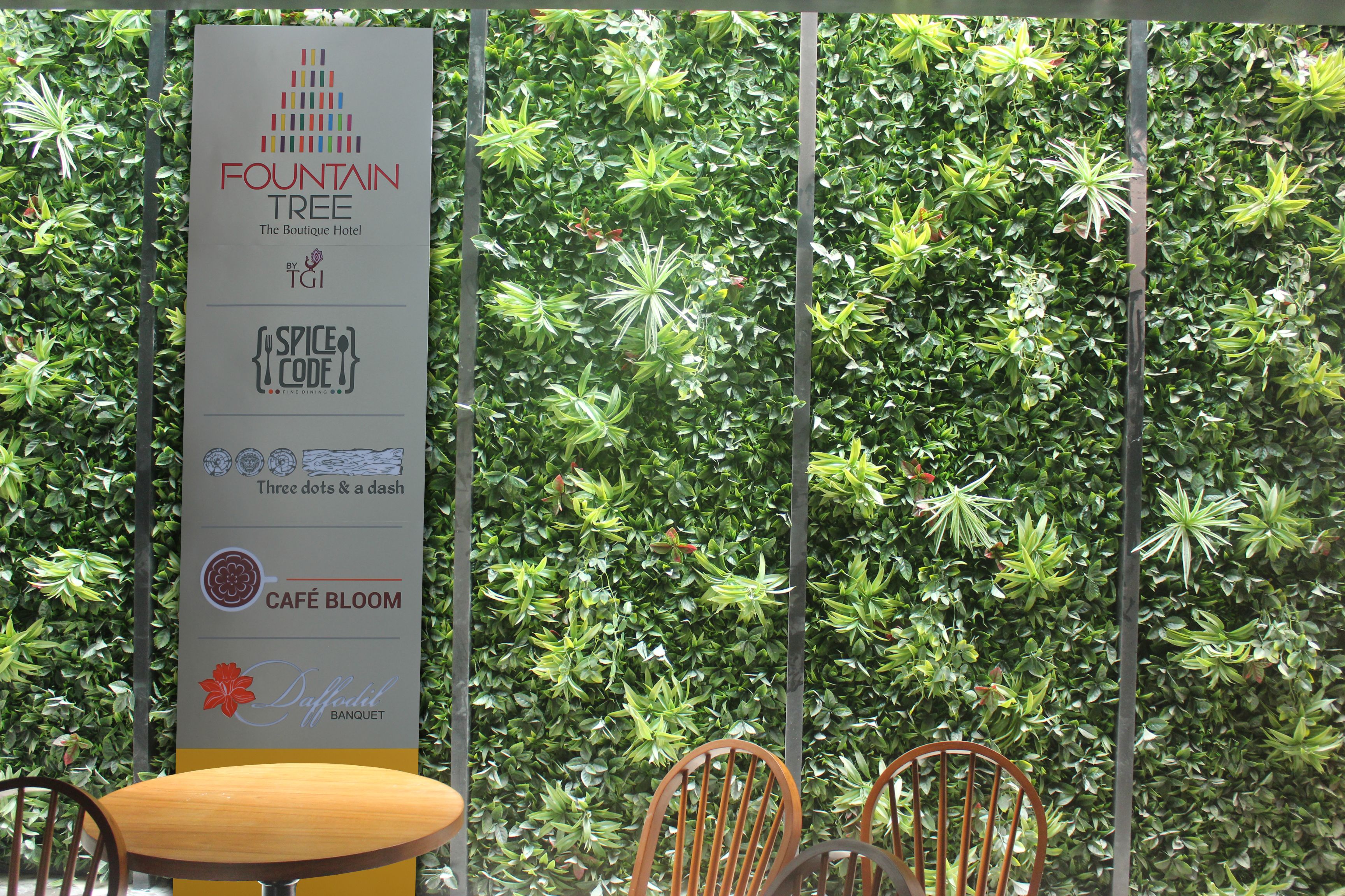 A vertical sign with logos and names of different hotel sections, including Fountain Tree, Spice Code, and Café Bloom, set against a backdrop of dense green foliage.