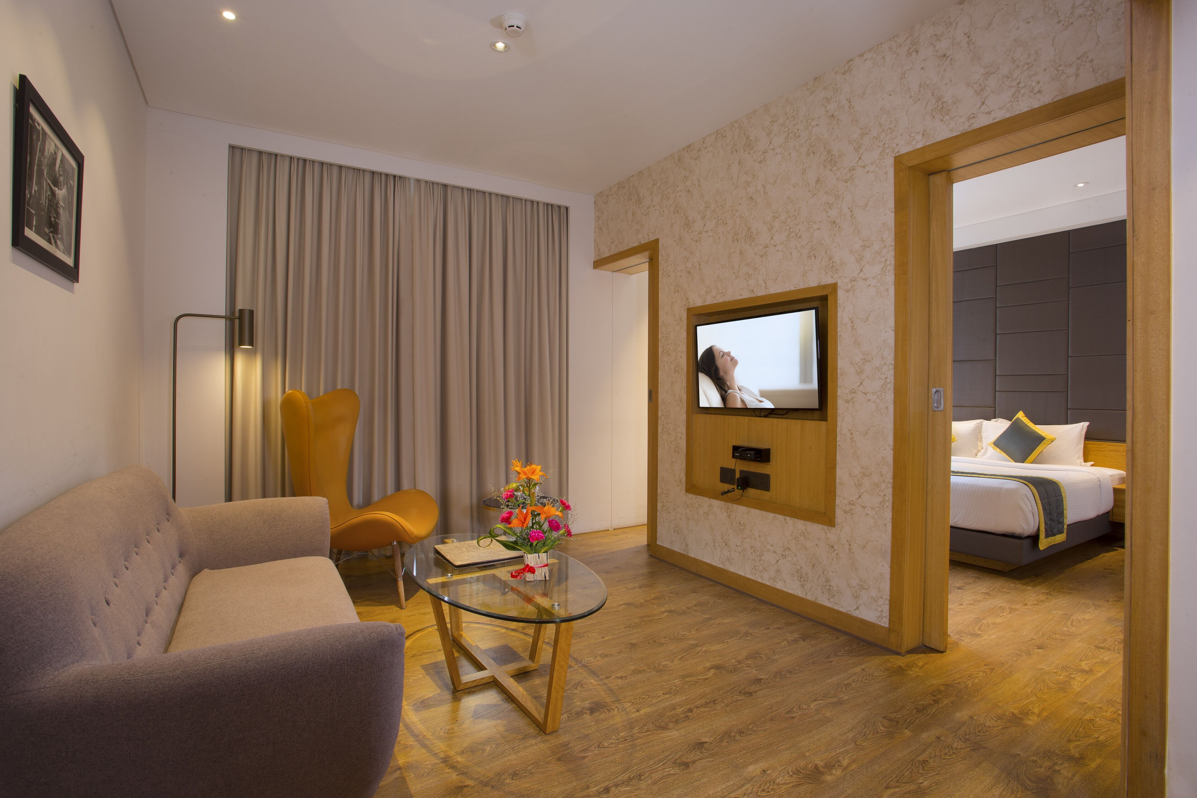 A modern hotel room with a sofa, a glass coffee table, a TV mounted on the wall, and a bed with a wooden headboard.