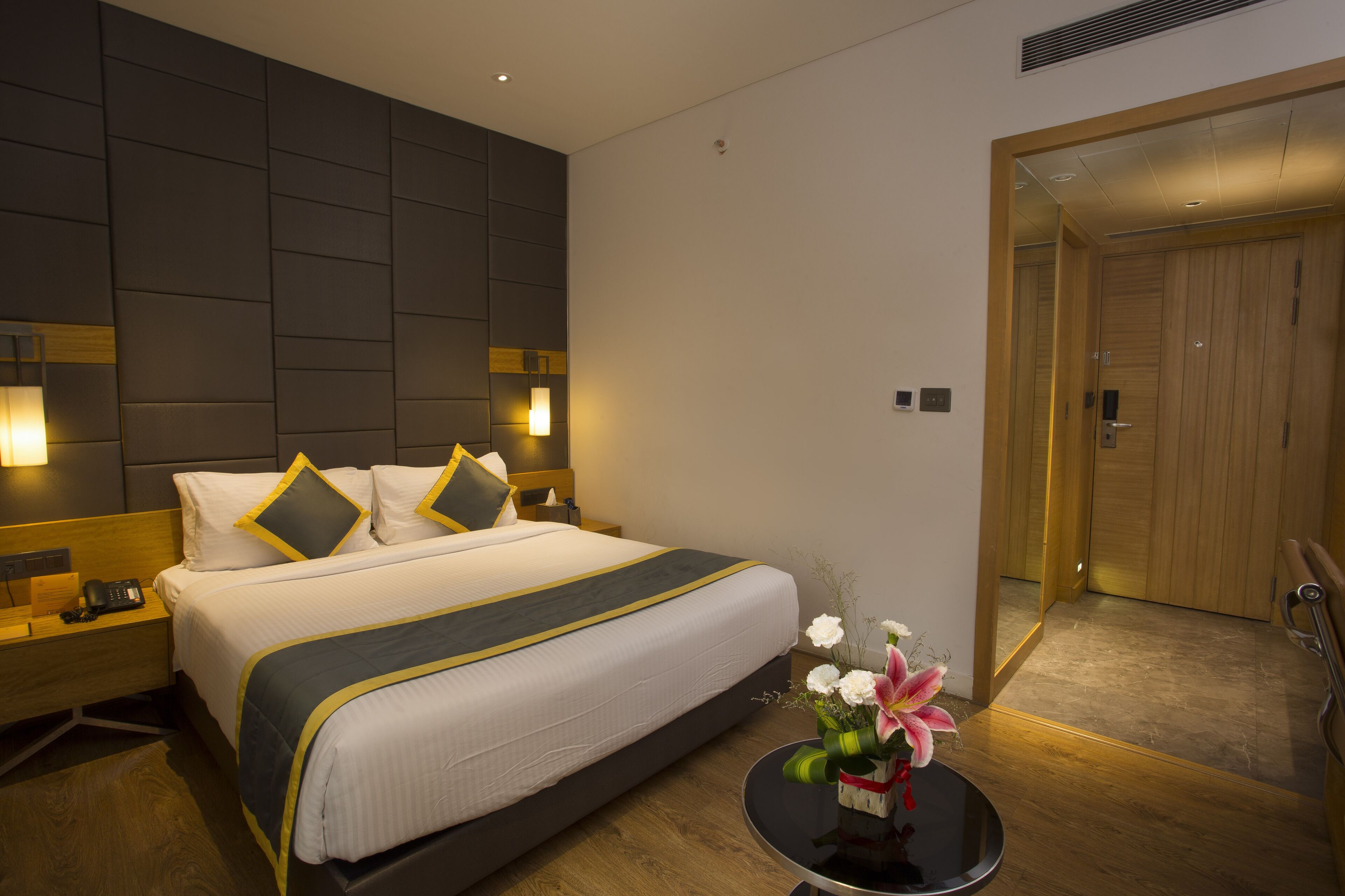 A hotel room with a large bed, bedside lamps, a bedside table, a small round table with a vase of flowers, and a wooden door.
